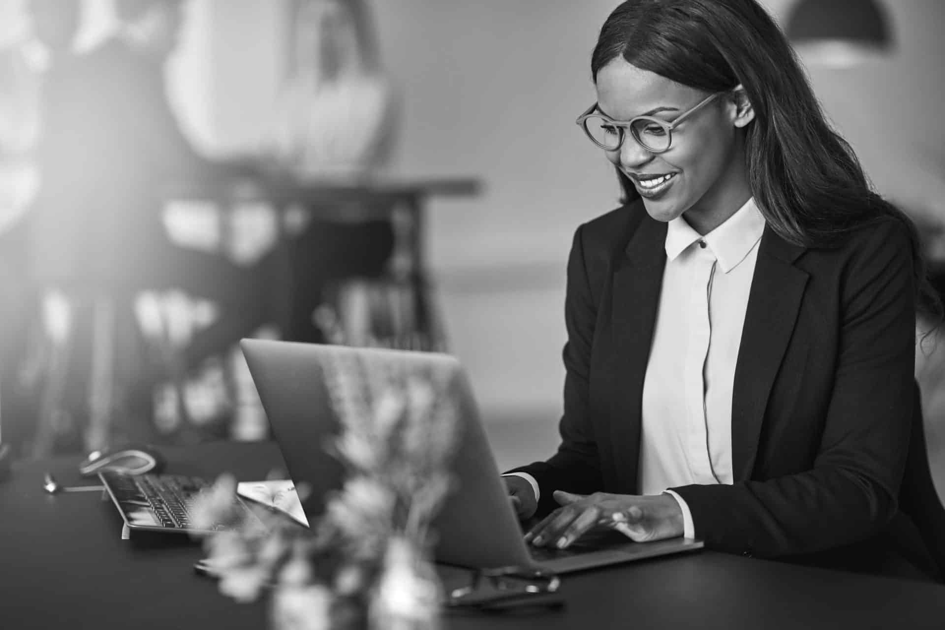 Smiling young African American businesswoman working on a laptop at her desk in a bright modern office with colleagues in the background