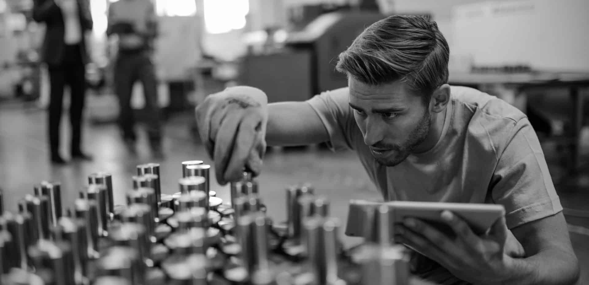 Young steel worker examining manufactured rod cylinder while using digital tablet in distribution warehouse.